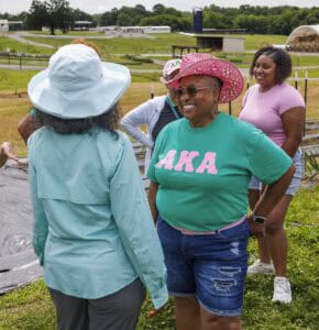 A smiling woman in a pink cowboy hat and green AKA shirt talks with another woman in a wide-brim hat in a sunny outdoor farm setting, while others stand and chat in the background.
