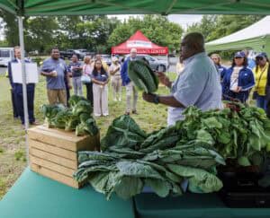 A man holds up a large leafy vegetable while speaking to a group of people gathered under outdoor tents at a community event. Tables display more fresh greens, and a red Granville County Center tent is visible in the background.