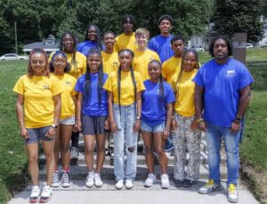 A group of fourteen people, mostly teens, pose outdoors on a sidewalk. They wear matching yellow and blue shirts, with some wearing shorts or jeans, standing in front of grass and trees on a sunny day.