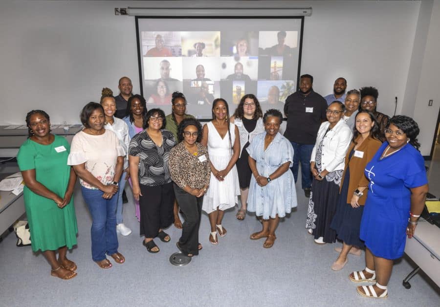 A group of people stand together in a classroom, smiling at the camera. A projector screen behind them shows a video call with several more participants, indicating a hybrid in-person and virtual meeting or event.