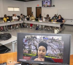 A classroom of people sit around tables while a computer monitor in the foreground shows a woman participating in a video call, with a tree-lined background on her screen.