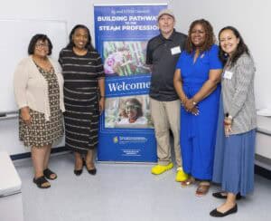 Five adults stand smiling beside a “Building Pathways to the STEAM Professions” welcome banner in a classroom, dressed in business casual attire. The group is diverse in gender and attire.