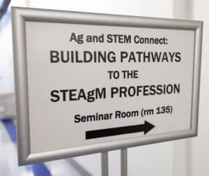 A sign reads, “Ag and STEM Connect: BUILDING PATHWAYS TO THE STEAgM PROFESSION. Seminar Room (rm 135)” with a black arrow pointing right. The sign is in a hallway with a blue floor visible on the left.