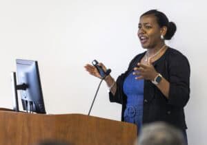A woman stands at a podium speaking, gesturing with her hands. She is wearing a blue dress and black jacket, and holding a presentation remote. A computer monitor and microphone are on the podium.