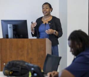A woman stands at a podium giving a presentation, holding a remote. She faces an audience as a man sits in the foreground taking notes. A computer monitor and backpack are visible.