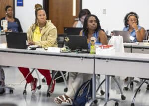 A group of women sit at tables with laptops and notebooks, attentively listening during a classroom or workshop setting. Some have snacks and drinks, and one woman wears bright red shoes.