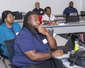 Several adults sit attentively in a classroom, facing forward. Some have laptops and drinks on the tables in front of them. The atmosphere appears focused and engaged.