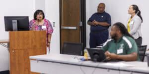 A woman speaks at a podium in a classroom while three adults—two standing, one seated at a table—listen attentively. The setting appears to be a professional or educational presentation.