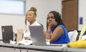 Two women sit at a desk with laptops in front of them, attentively listening in a classroom or meeting setting. One woman wears a blue top and has her chin in her hand, while the other wears a light-colored shirt.
