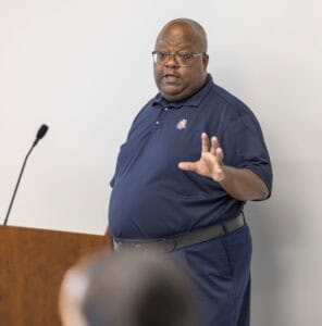 A man wearing glasses and a navy blue polo shirt stands near a podium, gesturing with one hand as he speaks, with a microphone stand visible in the background.