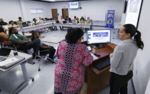 Two women stand at a podium presenting to an audience seated in a U-shaped arrangement in a classroom. A presentation is displayed on the monitor, and participants listen attentively.