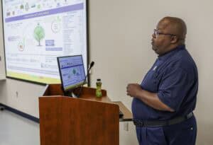 A man stands at a podium giving a presentation, with a slide projected on the wall and a computer monitor in front of him. He appears to be explaining data or a diagram.