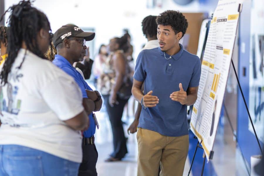 A young man speaks animatedly while presenting a poster to two adults in a busy hallway filled with other people and displays.