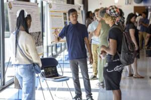 A young man explains his research poster to two students in a bright hallway during a poster presentation event, with other people and posters visible in the background.