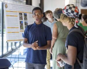 A young man in a navy polo shirt speaks enthusiastically to another person at a science fair or academic event, standing in front of a research poster. Other attendees mingle in the background.