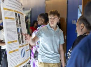 A young person in a light blue polo shirt points to information on a large poster board, presenting research to an audience in a hallway with several people in the background.