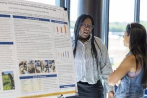 A woman in glasses and a striped shirt discusses a scientific research poster with another woman at an indoor event, with sunlight coming through large windows in the background.