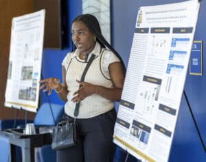 A young woman presents research displayed on a poster board, gesturing with her hands while speaking. Two research posters with charts and images are visible beside her in a conference setting.