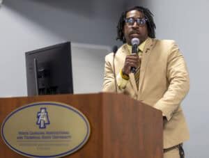 A person in a tan suit speaks into a microphone at a podium with a North Carolina Agricultural and Technical State University sign on it, in a conference or classroom setting.