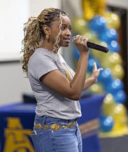 A woman with braided hair speaks into a microphone. She wears a gray t-shirt, jeans, and a gold belt. Blue and yellow balloons and a blue tablecloth are visible in the blurred background.