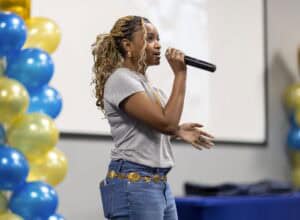 A woman with braided hair speaks into a microphone at an event, standing near blue and yellow balloon decorations. She wears a gray T-shirt, blue jeans, and a decorative belt.