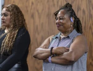Two women stand indoors against a wooden wall. The woman in front smiles with her arms crossed, wearing a sleeveless shirt and hearing aid. The woman behind her has long curly hair and wears a black top.