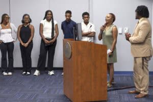 Six young adults stand in a line, while a woman speaks into a microphone next to a man in a suit by a wooden podium in a conference room.