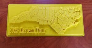 A yellow 3D-printed plaque shows a map of North Carolina with county borders and the text 2025 Eastern Tour on a reddish wood surface.