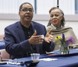 A man in a navy suit gestures while speaking at a table, with a woman in a tan shawl sitting beside him. A small vase with flowers and a cup are on the table in front of them.