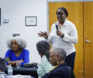 A woman stands and speaks into a microphone in a meeting room, while several older adults sit and listen at tables. A wooden door and framed items are visible on the wall in the background.