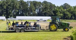 A green tractor pulls an open-air trailer with seated people on a grassy field; solar panels, a pickup truck, and trees are visible in the background under a clear blue sky.