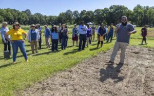 A man speaks to a diverse group of people gathered outdoors in a grassy field on a sunny day, with trees in the background. The group listens attentively, some taking notes and others smiling.
