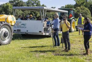 A group of people attend an outdoor agricultural event; some sit in a covered trailer, while three people, one speaking into a microphone, stand in front of a tractor on a sunny day.