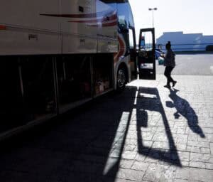 A person walks toward the open door of a parked tour bus, casting a long shadow on the pavement in bright sunlight. The bus storage compartments are open, revealing luggage inside.