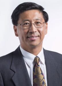 A man wearing glasses, a dark suit, a white shirt, and a colorful floral-patterned tie, smiling, with short dark hair, posing against a plain light background.