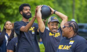 Three people in NC A&T shirts participate in an outdoor group activity, smiling and passing a large black ball over their heads, with greenery in the background.
