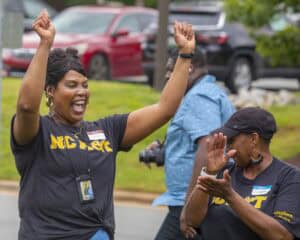 Two women outdoors celebrate joyfully; one raises her arms and smiles widely while the other claps. Both wear black NCAT shirts and name tags, with cars and greenery visible in the background.