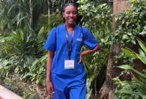 A woman in blue medical scrubs with an ID badge stands outdoors, smiling in front of lush green plants and trees.