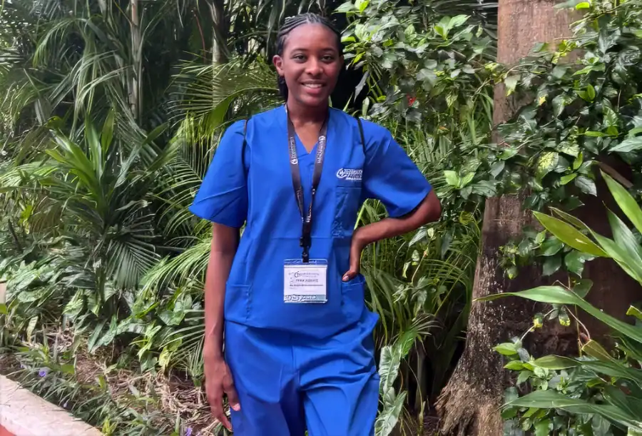 A woman in blue medical scrubs with an ID badge stands outdoors, smiling in front of lush green plants and trees.