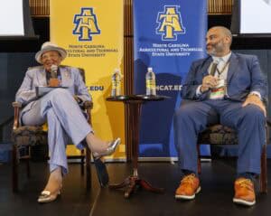 Two people sit on stage in conversation, each holding a microphone, in front of North Carolina A&T State University banners. Both wear suits; the woman on the left smiles, wearing a hat, while the man on the right listens attentively.