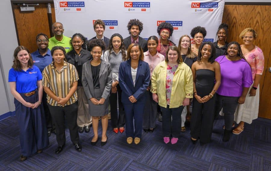 A diverse group of 20 people stand and smile for a group photo in front of an NC Cooperative Extension banner in an indoor setting.