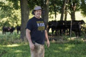 A man wearing a wide-brimmed hat and a t-shirt stands smiling in a grassy field with a group of black cows and trees in the background on a sunny day.