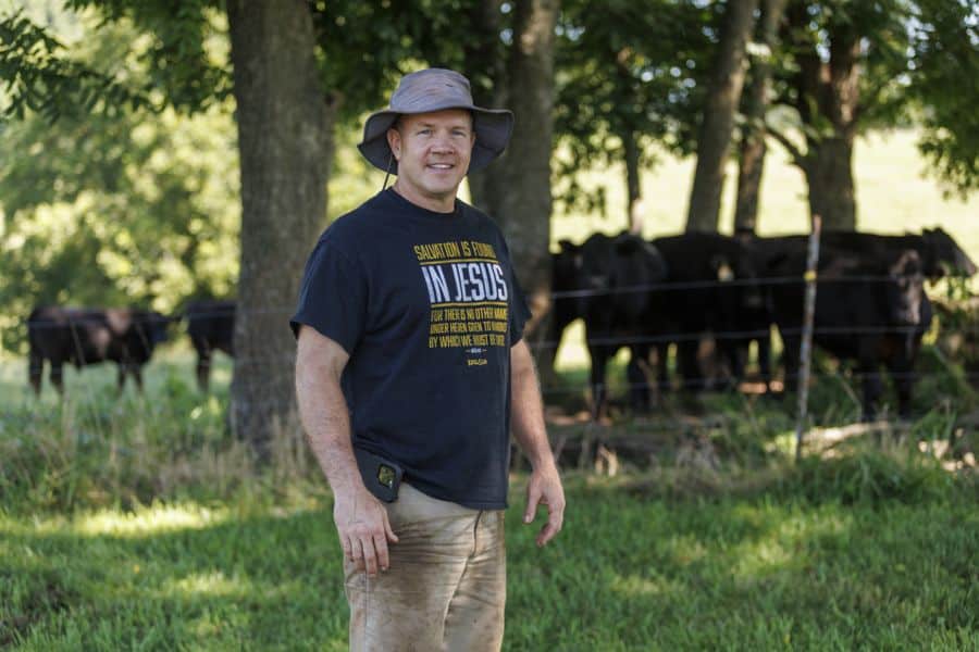 A man wearing a wide-brimmed hat and a t-shirt stands smiling in a grassy field with a group of black cows and trees in the background on a sunny day.