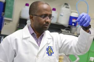 A scientist in a white lab coat with an A&T logo examines a test tube in a laboratory, wearing blue gloves and glasses, with lab equipment and bottles in the background.