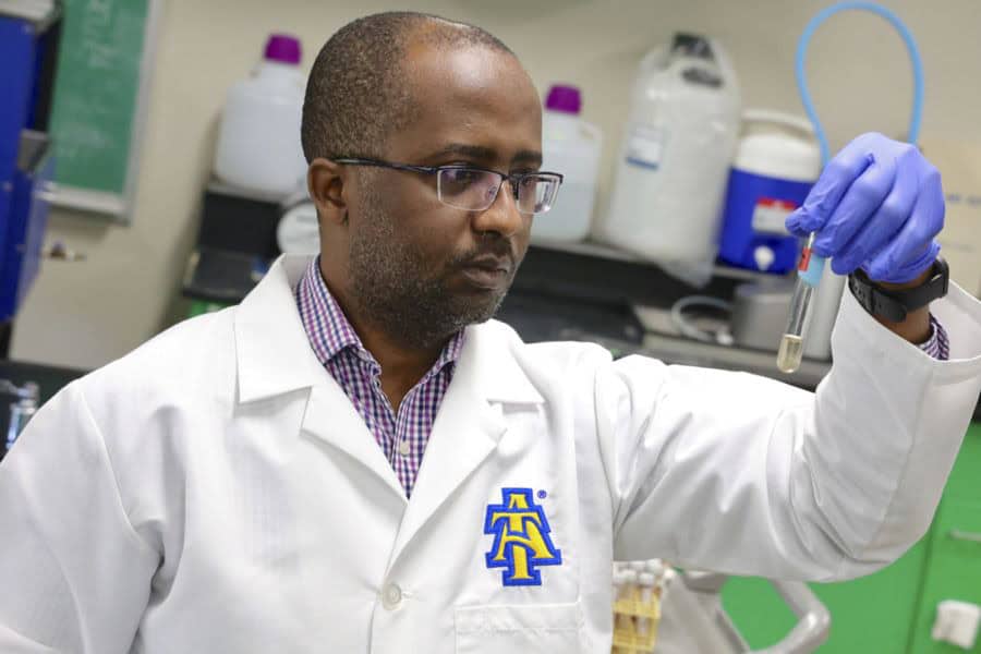 A scientist in a white lab coat with an A&T logo examines a test tube in a laboratory, wearing blue gloves and glasses, with lab equipment and bottles in the background.