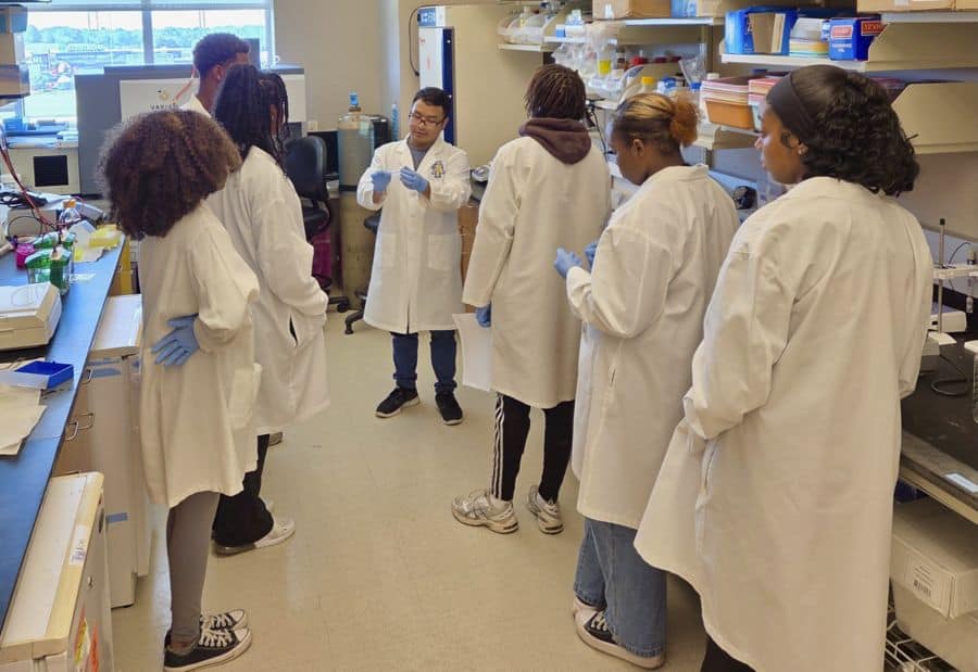 A group of students in white lab coats and blue gloves listen to an instructor explaining something in a science laboratory filled with equipment and supplies.