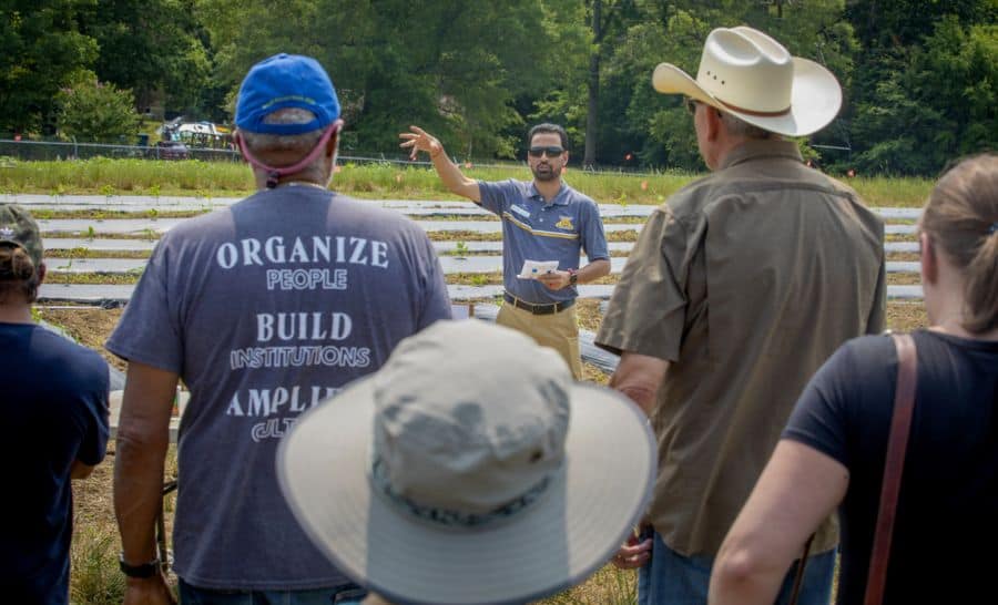 A man speaks to a group of people outdoors in front of garden beds. The group listens attentively, some wearing hats. Trees and greenery are visible in the background. One person’s shirt reads “ORGANIZE PEOPLE BUILD INSTITUTIONS AMPLIFY CULTURE.”.