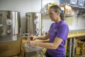 A woman in a purple shirt fills small glass bottles with liquid in a workspace, standing next to large stainless steel tanks and shelves with supplies.