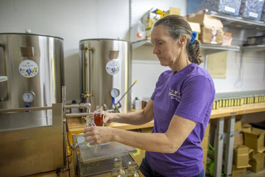 A woman in a purple shirt fills small glass bottles with liquid in a workspace, standing next to large stainless steel tanks and shelves with supplies.
