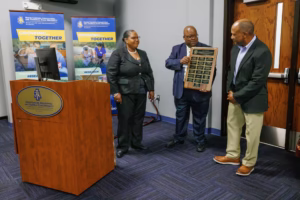 Three people stand in a room; one man holds a plaque while another man and a woman look on. Behind them are banners reading Growing... Together and a podium with an NC A&T State University logo.